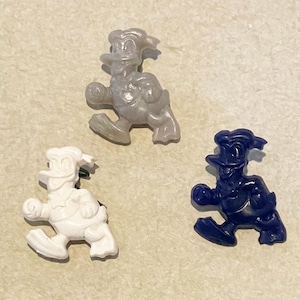 Könnte beinhalten: Drei Plastikfiguren eines Comic-Entchens in verschiedenen Farben: grau, weiß und blau. Das Entchen trägt einen Hut und rennt.