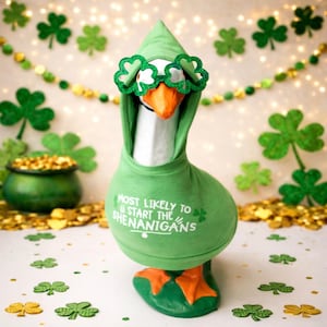 May include: A goose figurine dressed in a green hoodie with the text "MOST LIKELY TO START THE SHENANIGANS". The goose wears shamrock-shaped glasses and has orange feet. The background includes shamrock decorations and a pot of gold.