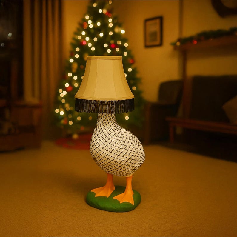 Op de afbeelding: Een grillige lamp in de vorm van een gans, met een beige lampenkap en zwarte franjes. De gans heeft een wit lichaam met een zwart netpatroon, oranje voeten en staat op een groene basis. Op de achtergrond is een kerstboom te zien.