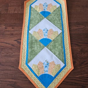 May include: A handmade, quilted table runner with a geometric design. The runner features a central pattern of green, white, and blue shapes, bordered by yellow floral fabric and an orange edge. The runner is placed on a wooden surface.