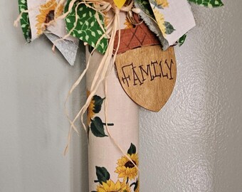 Op de afbeelding: Een decoratieve deegroller met een crèmekleurige body met zonnebloemprint, bekroond met een grote gele zonnebloem en stoffen linten. Een houten eikelvormig label hangt bovenaan met het woord FAMILY erin gebrand.