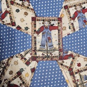 Op de afbeelding: Een patchwork quiltstof met een herhalend patroon van vogelverschrikkers in blauwe tuinbroeken en rode overhemden, afgewisseld met blauwe stoffen panelen bedrukt met kleine witte hartjes. Een centrale vogelverschrikkerpatch toont de tekst GOOD HARVEST.