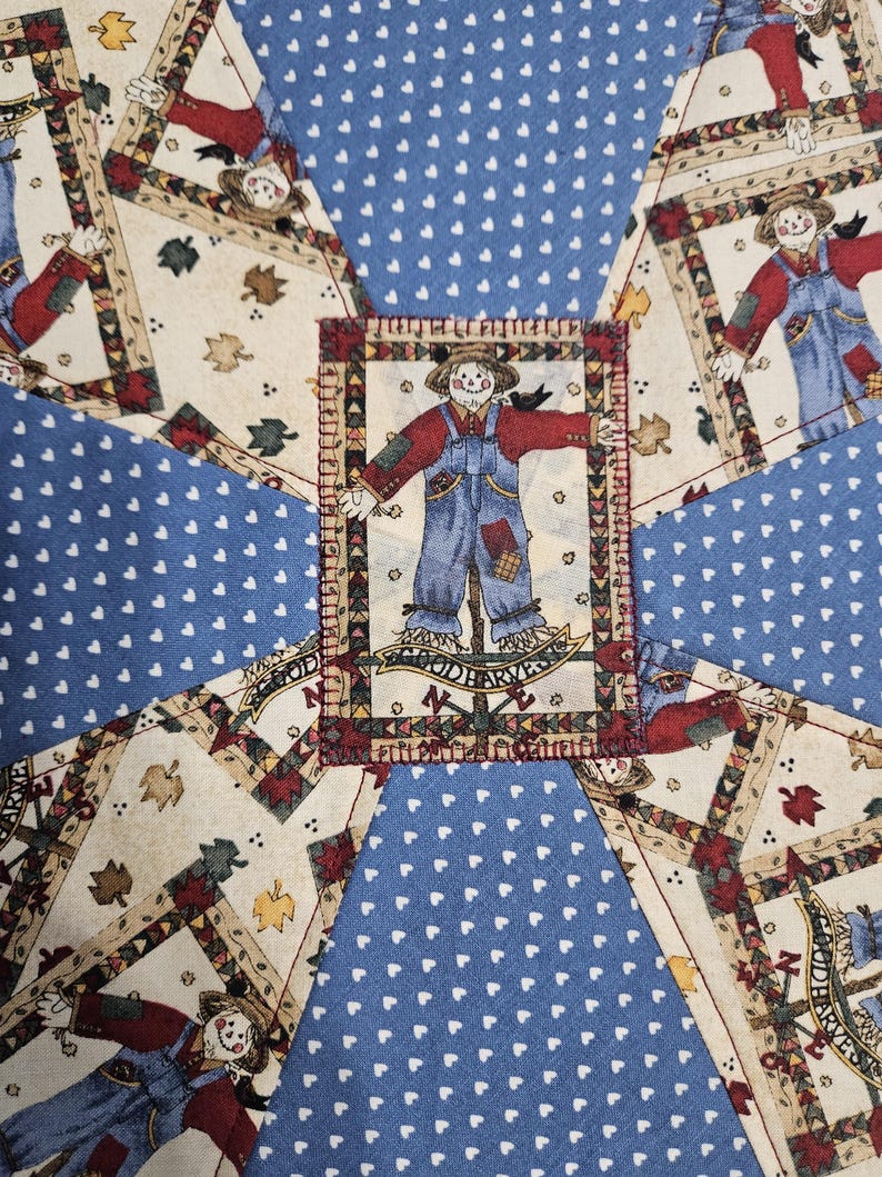 Op de afbeelding: Een patchwork quiltstof met een herhalend patroon van vogelverschrikkers in blauwe tuinbroeken en rode overhemden, afgewisseld met blauwe stoffen panelen bedrukt met kleine witte hartjes. Een centrale vogelverschrikkerpatch toont de tekst GOOD HARVEST.