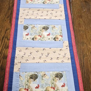 May include: A rectangular table runner with a rooster and butterfly pattern. The runner features alternating light blue and beige fabric sections, with a red border and blue trim. The rooster pattern is on a light blue background.