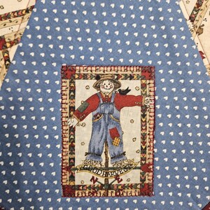 Op de afbeelding: Een blauw stoffen paneel met een wit hartjespatroon en een centrale rechthoekige patch. De patch toont een vogelverschrikker in een rood hemd en blauwe tuinbroek, met de tekst "GOOD HARVEST" eronder, omlijst door een kleurrijke driehoekige rand.