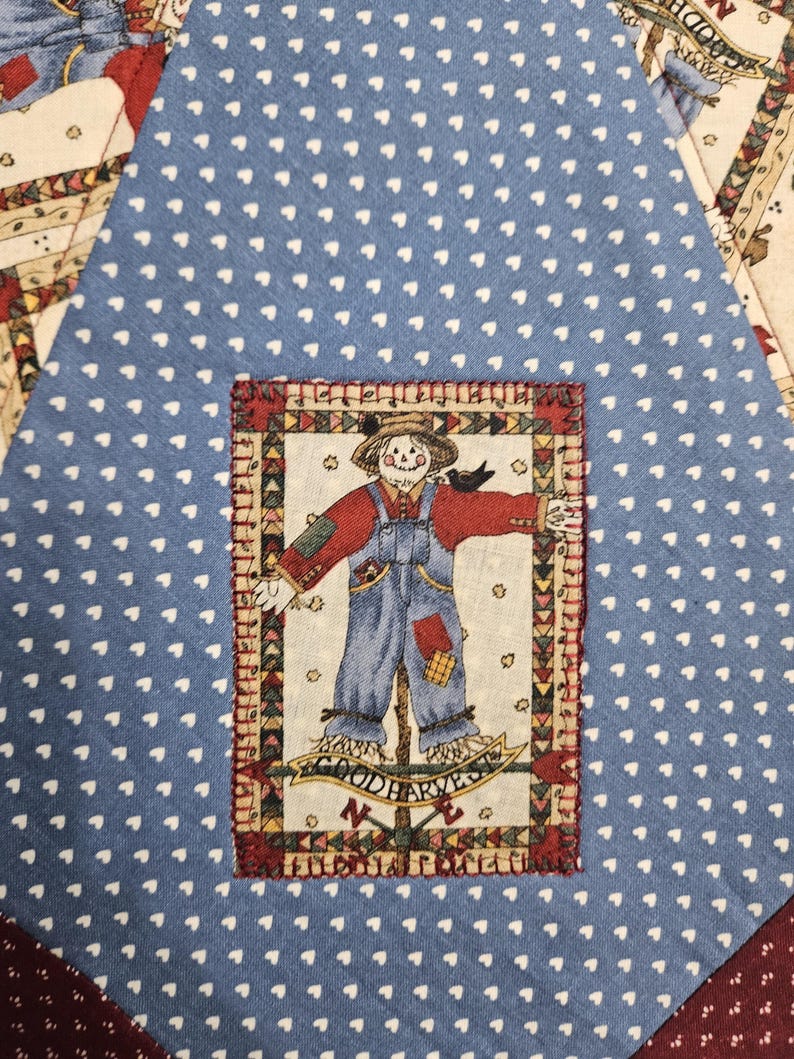 Op de afbeelding: Een blauw stoffen paneel met een wit hartjespatroon en een centrale rechthoekige patch. De patch toont een vogelverschrikker in een rood hemd en blauwe tuinbroek, met de tekst "GOOD HARVEST" eronder, omlijst door een kleurrijke driehoekige rand.