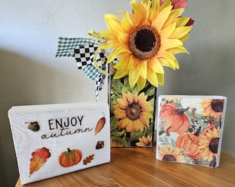 Op de afbeelding: Een rechthoekig wit houten blok met de tekst ENJOY autumn, versierd met pompoen- en bladstickers. Daarnaast staat een hoge rechthoekige vaas met zonnebloemafbeeldingen en een zwart-wit geblokt lint, gevuld met kunstmatige zonnebloemen.