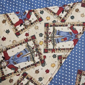 Op de afbeelding: Een stof met een herhalend patroon van vogelverschrikkers in blauwe tuinbroeken en rode shirts, omlijst door decoratieve randen, afgewisseld met herfstbladeren op een cr&egrave;mekleurige achtergrond, gecontrasteerd met delen blauwe stof met witte hartjes.