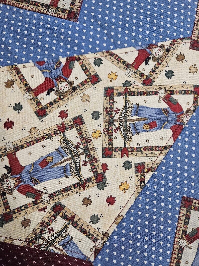 Op de afbeelding: Een stof met een herhalend patroon van vogelverschrikkers in blauwe tuinbroeken en rode shirts, omlijst door decoratieve randen, afgewisseld met herfstbladeren op een cr&egrave;mekleurige achtergrond, gecontrasteerd met delen blauwe stof met witte hartjes.