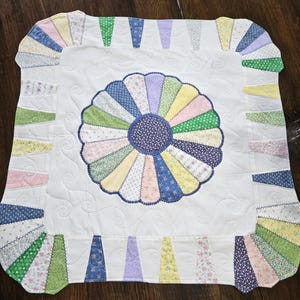 May include: A handmade, square quilt featuring a large, colorful flower design in the center. The flower is surrounded by a white background with a scalloped edge. The quilt is made of various patterned fabrics in pastel colors.