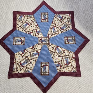 May include: A star-shaped quilted table topper featuring alternating panels of light blue fabric with white polka dots and a cream-colored fabric printed with scarecrows and autumn leaves, all bordered by a dark red fabric with white dots.