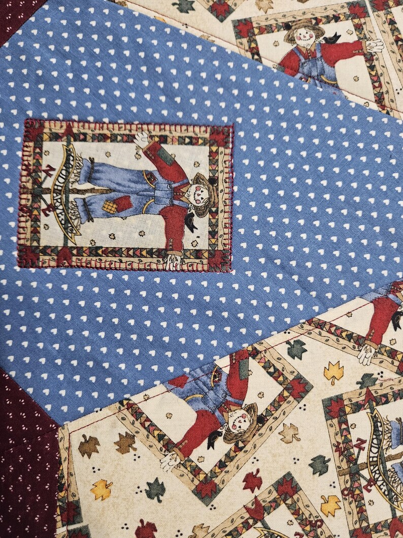 Op de afbeelding: Een patchwork quilt met blauwe stof met een wit hartjespatroon, cr&egrave;mekleurige stof met herfstbladmotieven en donkerbordeauxrode stof met witte stippen. Rechthoekige patches tonen vogelverschrikkers met de tekst "HARVEST".