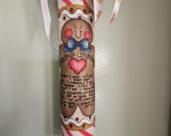 Op de afbeelding: Een decoratieve deegroller met een illustratie van een peperkoekmannetje met een blauwe strik en een rood hart. De tekst luidt Grandma's Gingerbread en bevat een recept. De uiteinden zijn beschilderd met rood-witte glitterstrepen.