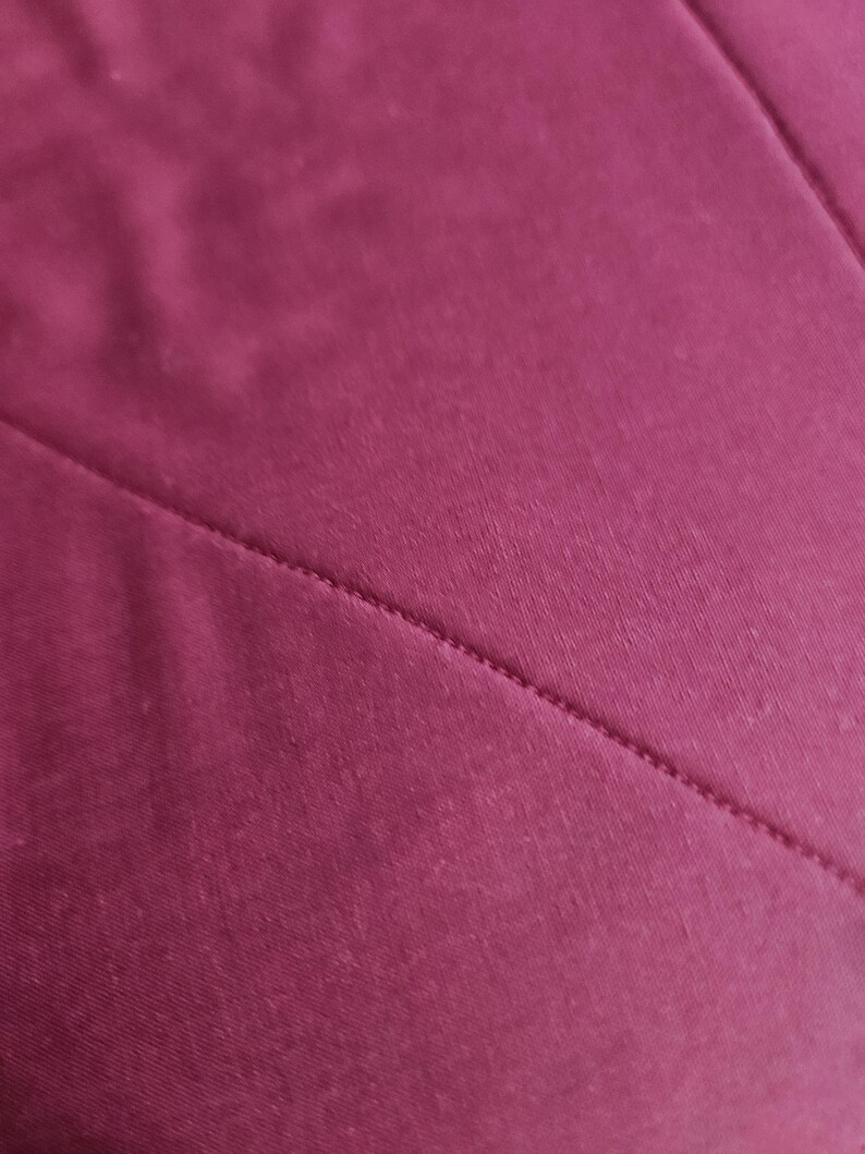 Op de afbeelding: Een effen diep magenta stof met een diagonale naad die over het oppervlak loopt. Het materiaal heeft een gladde, matte textuur met een consistente kleur en toont een strakke stiklijn waar twee delen van het textiel samenkomen.