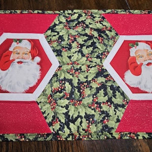 May include: A quilted table runner featuring a repeating pattern of Santa Claus portraits in red and white, alternating with hexagonal sections of green fabric decorated with holly leaves and red berries, all bordered by a red glittery fabric trim.