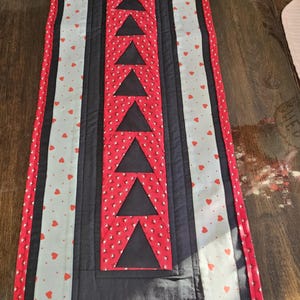 May include: A rectangular quilted table runner with a central black panel featuring red triangles. The runner has a red border and alternating panels of red heart-patterned fabric and solid black fabric. A decorative home accent.