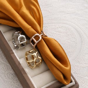 May include: Three metal napkin rings in silver, rose gold, and gold tones, each featuring a geometric open-link design. One ring is threaded onto a folded mustard yellow satin fabric napkin, while the other two rest in a rectangular display box.