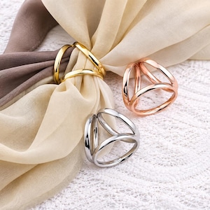 May include: Three circular scarf rings in gold, rose gold, and silver tones, each featuring a triple-loop design. One gold ring secures a beige and brown fabric scarf, while the rose gold and silver rings are positioned separately nearby.