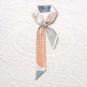 May include: A long, narrow neck scarf featuring a patchwork design with various patterns including floral motifs, geometric grids, and polka dots in shades of peach, light blue, white, and red. The scarf is tied in a simple knot at the top.