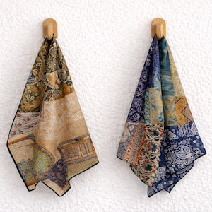 May include: Two decorative scarves hanging on wooden hooks against a white textured wall. The scarves feature a patchwork design with various patterns and colors, including beige, blue, and gold. The scarves are folded and draped.