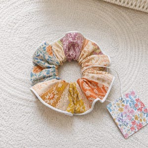 May include: A fabric hair scrunchie featuring a patchwork of floral and botanical patterns in shades of orange, yellow, blue, and purple, finished with a white piped edge and a square tag displaying a colorful wildflower print.