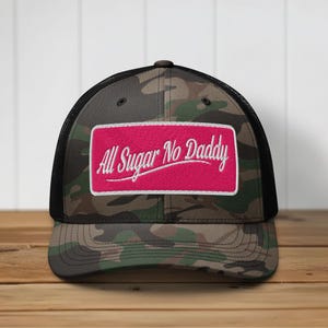 May include: A camouflage print baseball cap with a black mesh back and a rectangular pink patch on the front. The patch features the white embroidered text All Sugar No Daddy in a cursive script font with a white border around the patch edge.