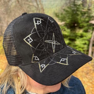 May include: Black trucker cap with a geometric design in silver and gold. The cap has a mesh back and a curved peak. The design includes a central star and angular shapes, creating a unique accessory.