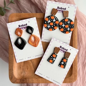 May include: Three pairs of earrings on display cards labeled Baked Design. One pair features black and orange glittery diamond shapes. Another pair has round black discs with orange pumpkins and white stars. The third pair has black trapezoids with pumpkins.