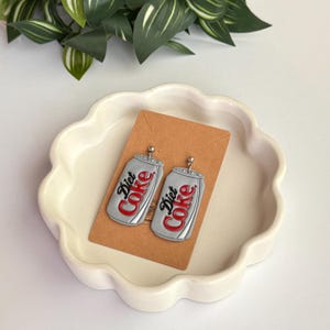 May include: A pair of Diet Coke can earrings. The earrings are silver with red and black text that reads "Diet Coke". The earrings are displayed on a brown card in a white, scalloped dish.