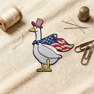 May include: Embroidered patch of a white goose wearing a patriotic hat and cape with the American flag design. The goose has a yellow beak and feet. Other sewing supplies are in the background.