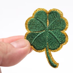 May include: Embroidered four-leaf clover patch with a green clover and a gold glitter border. The patch is held up against a white background. This patch is perfect for St. Patrick's Day or any occasion.