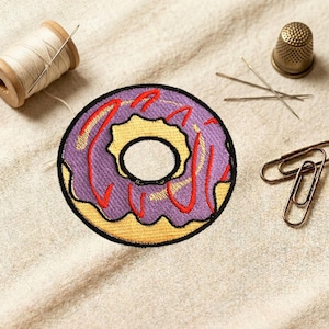 May include: Embroidered donut patch with purple frosting and red drizzle. The patch has a yellow center and a black outline. A wooden spool of thread, needles, a thimble, and paper clips are also in the image.