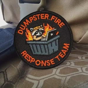 May include: Round black patch with orange text that reads "DUMPSTER FIRE RESPONSE TEAM." The patch features a raccoon in a hard hat working on a laptop in a dumpster engulfed in flames.