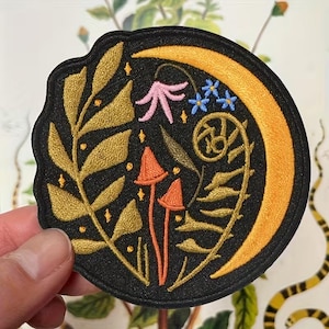 May include: Embroidered patch with a black background featuring a gold crescent moon, gold leaves, orange mushrooms, pink flower, and blue flowers. The patch has a unique, irregular shape and is likely for clothing or accessories.