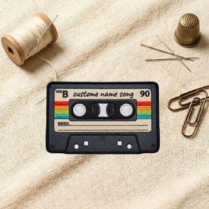 May include: Embroidered patch in the shape of a cassette tape with the text "custom name song 90". The patch has a rainbow stripe design and is black, beige, and white. A spool of thread, needles, a thimble, and paper clips are also in the image.
