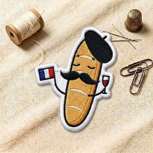 May include: Embroidered patch featuring a cartoon baguette wearing a beret and mustache, holding a French flag and a glass of wine. The patch is yellow, black, white, red, and blue. Other items in the image include a spool of thread, a thimble, needles, and paper clips.