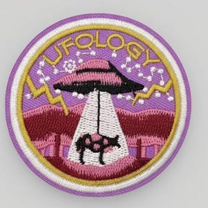May include: Embroidered patch with a purple background and the word "UFOLOGY" in gold. The patch features a black and pink UFO beaming a cow. The design includes pink and maroon mountains, and gold lightning bolts.