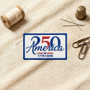 May include: A rectangular embroidered patch with a white background and a blue border. The design features the blue text America, a large red number 50, and the text 1776-2026 with a small blue star in the center, all stitched in red and blue thread.