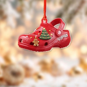 May include: A red Christmas ornament shaped like a Croc shoe, with the name "SOPHIA" on the side. The shoe is decorated with a green Christmas tree, a gingerbread man, and star-shaped accents. It hangs from a red string.