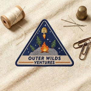 May include: A triangular embroidered patch featuring a dark blue night sky with stars, a campfire on a gray rock, green pine trees, and a small rocket ship. The text "OUTER WILDS VENTURES" is stitched in dark blue on a light beige horizontal band.