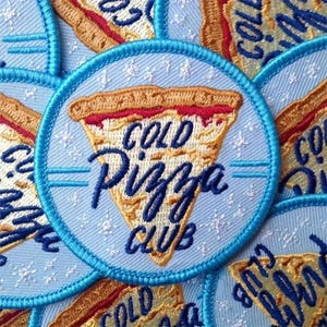 May include: Embroidered patch featuring a pizza slice with the text "COLD Pizza CLUB" in blue. The patch has a light blue background with a blue border and a pizza slice design in shades of brown, red, and yellow.