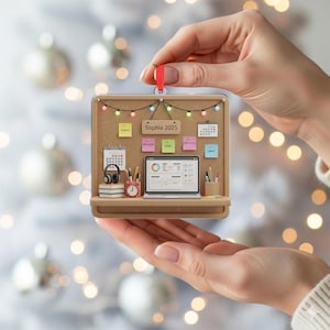 May include: A brown desk ornament with a corkboard background, featuring a miniature laptop, calendar, and headphones. The ornament has a red ribbon for hanging and includes the text "Sophia 2025".
