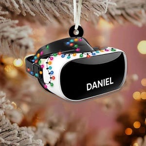 May include: A white and black VR headset Christmas ornament with the name "DANIEL" printed on the front. The ornament is decorated with colorful string lights and hangs from a white ribbon.