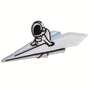 May include: Embroidered patch featuring a cartoon astronaut riding a stylized, silver rocket. The astronaut is depicted in black and white, with the rocket having a light blue accent. The patch is designed for application on clothing or accessories.