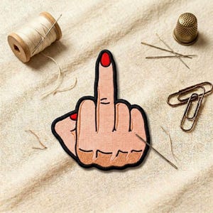 May include: Embroidered patch featuring a hand with the middle finger raised, outlined in black, with red nail polish. The patch is on a beige fabric, with sewing supplies like needles, thread, and a thimble scattered around.