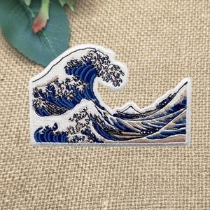May include: Embroidered patch featuring a stylized depiction of a large ocean wave in shades of blue, white, and tan. The design includes a mountain in the background, all outlined in white. The patch is likely for clothing or accessories.