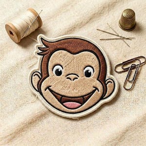 May include: Embroidered patch featuring a smiling monkey face with brown and tan thread. A spool of thread, needles, a thimble, and paper clips are also visible.