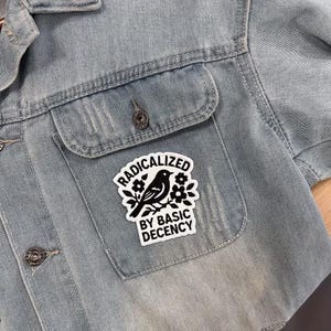 Pu&ograve; includere: Giacca di jeans azzurra con una toppa sulla tasca. La toppa presenta un uccello nero su un ramo con fiori e il testo "RADICALIZED BY BASIC DECENCY" in un bordo bianco. La giacca ha due tasche sul petto e bottoni in metallo.