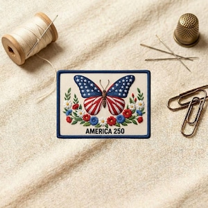 May include: A rectangular embroidered patch featuring a butterfly with wings patterned like the American flag, surrounded by red, white, and blue flowers. The text "AMERICA 250" is stitched in black at the bottom against a cream-colored background.