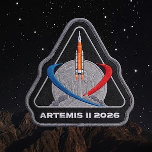 May include: A black and gray embroidered patch featuring the Artemis II 2026 mission logo. The design includes a rocket launching from a gray planet, with blue and red curved lines. The background shows a starry night sky and a brown mountain range.